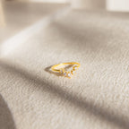The Marina Chevron Ring, a gold engagement ring adorned with small diamonds, rests on a textured beige surface in soft sunlight.