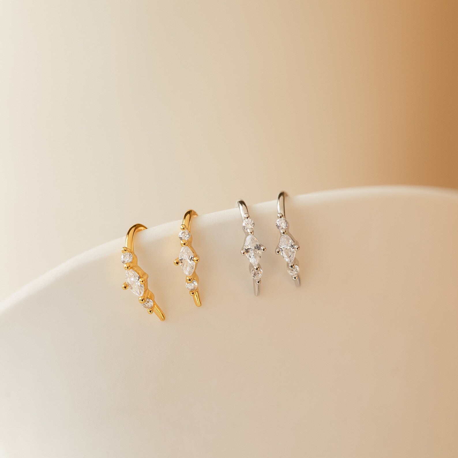 Two pairs of Amaya Marquise Threaders—one in gold, one in silver—adorned with dainty diamond clusters. These curved earrings sparkle beautifully displayed on a white surface.