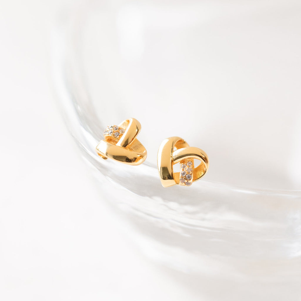 Heart Knot Studs adorned with small crystals feature a chic knot design and are elegantly displayed on a clear glass surface.
