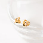 Heart Knot Studs adorned with small crystals feature a chic knot design and are elegantly displayed on a clear glass surface.