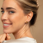 A woman smiles, showing off gold Heart Knot Studs with a delicate knot design. Her hair is styled back, and she wears a light-colored top.