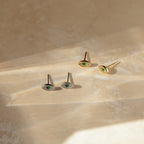 The Haven Emerald Eye Studs, a set of four elegant earrings with emerald stones, are showcased on a beige textured surface in gentle sunlight.