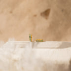 The Opal Inlay Huggies Set features two gold hoops with green opal accents, displayed on a light textured surface with a beige blurred background, highlighting their shimmering opal hues.
