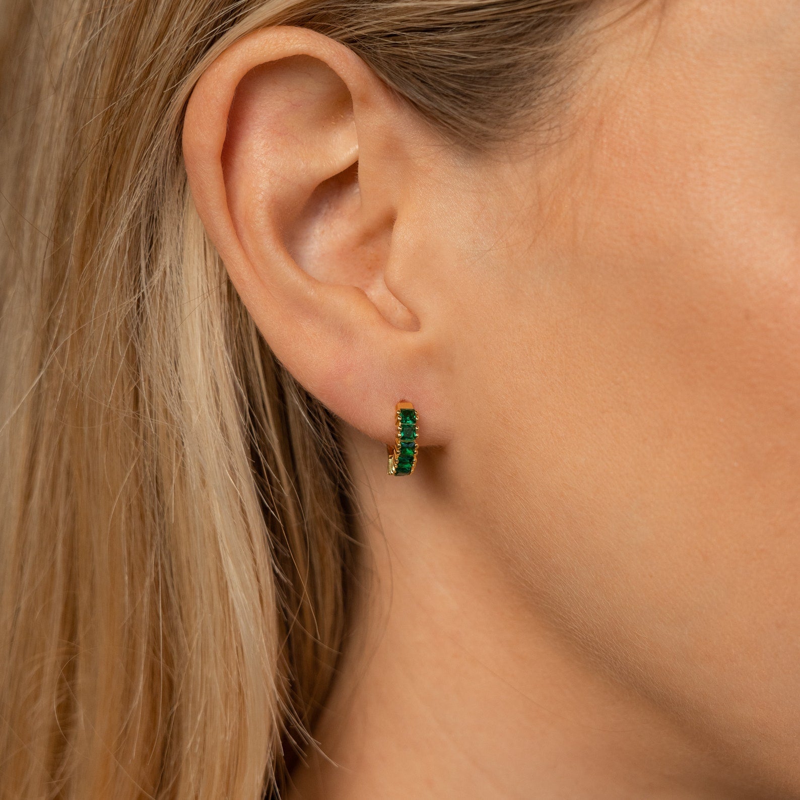 Close-up of a woman wearing the Genevieve Birthstone Huggie in emerald green, styled on her earlobe with blonde hair tucked behind her ear.