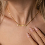 The Casella Name Necklace in Box Chain features a dainty script spelling Brooklyn, worn by a woman with light skin and manicured nails—an ideal personalized gift for any occasion.