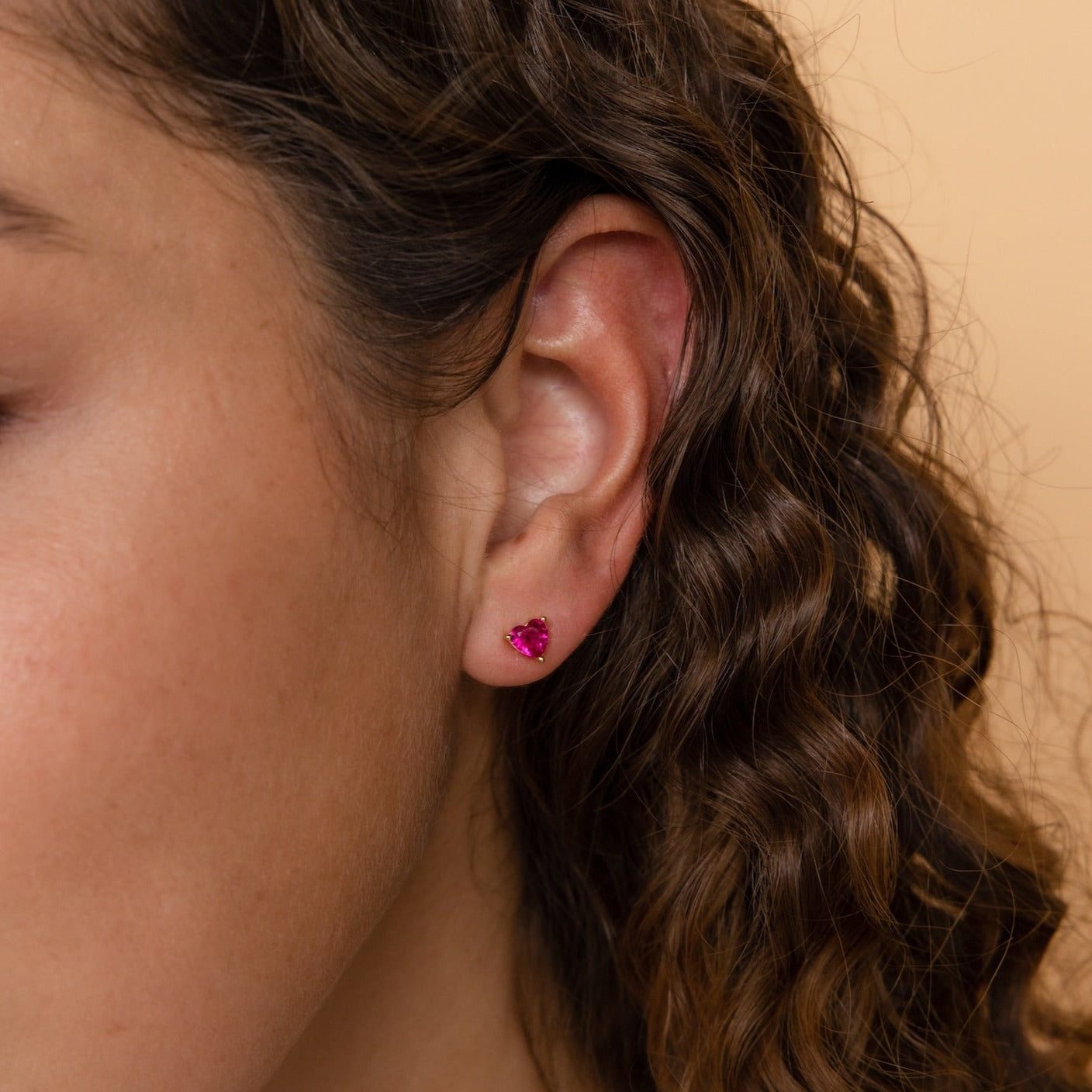 Close-up of a woman's ear with curly brown hair, wearing Ruby Heart Studs—small, heart-shaped earrings perfect for adding sparkle to your date night jewelry collection.