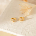 The Cobra Snake Huggies, gold rings featuring small green and clear gemstones, are perfect for fans of trendy jewelry and are displayed on a textured white surface near dried flowers.