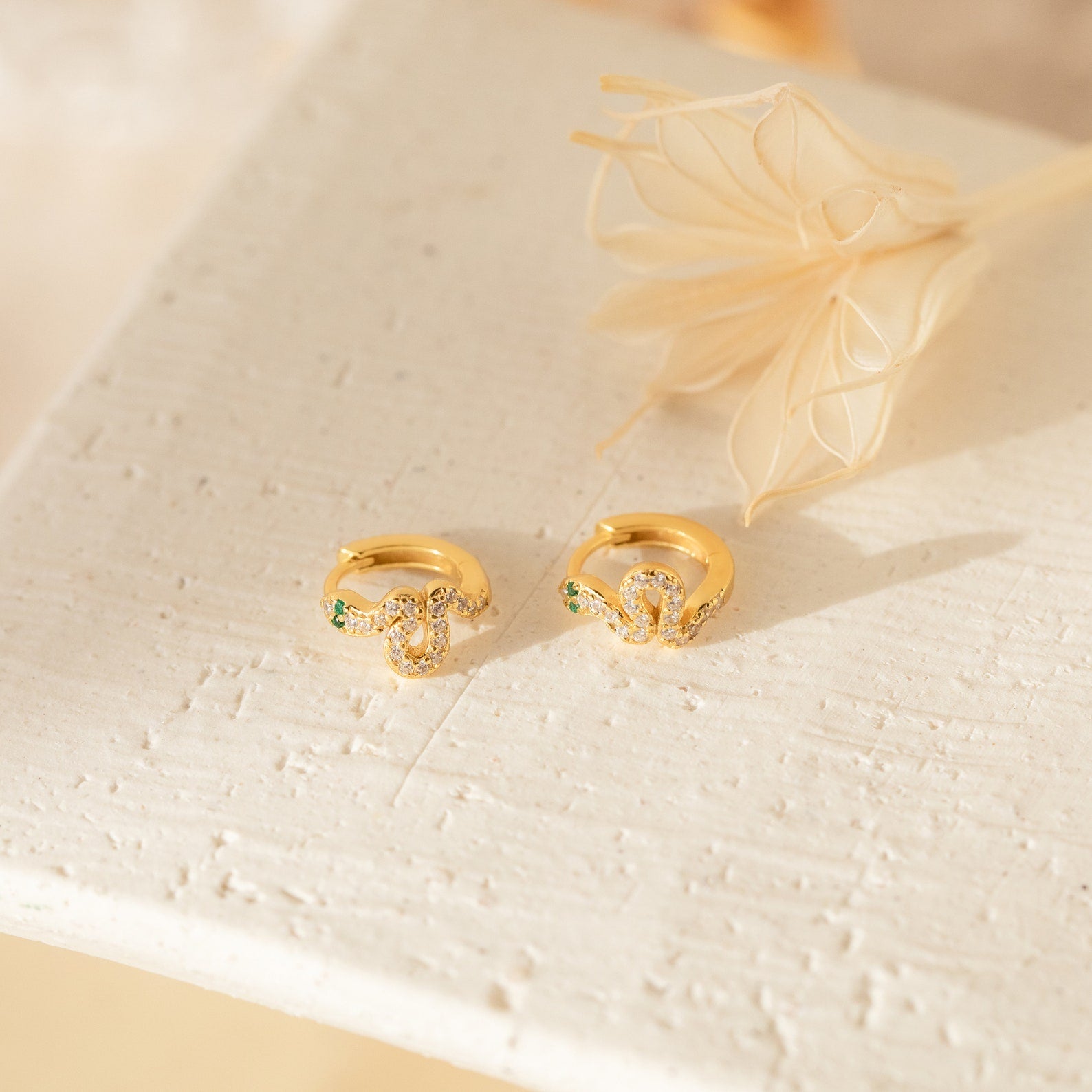 The Cobra Snake Huggies, gold rings featuring small green and clear gemstones, are perfect for fans of trendy jewelry and are displayed on a textured white surface near dried flowers.