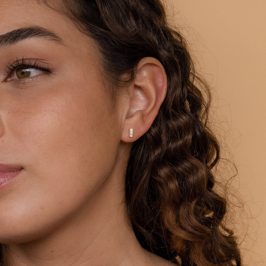 Close-up of a woman with curly hair wearing Serena Baguette Studs, minimal textured gold stud earrings, against a beige background.