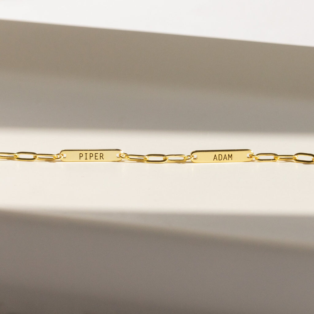 The Mini Name Bar Necklace features two rectangular name plates engraved with PIPER and ADAM, evoking the style of a personalized bar necklace, displayed against a neutral background.