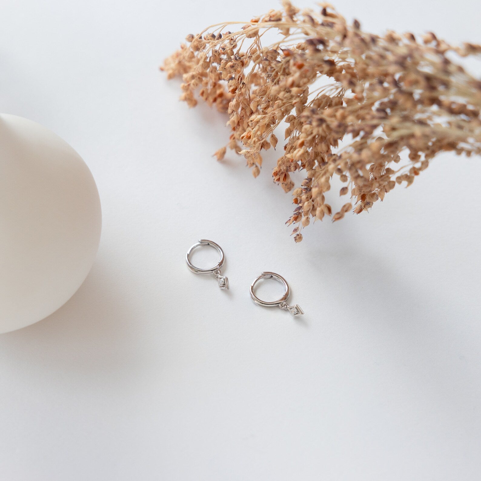 The Ericka Diamond Huggies in Sterling Silver are displayed on a white surface beside dried flowers and a round white object.