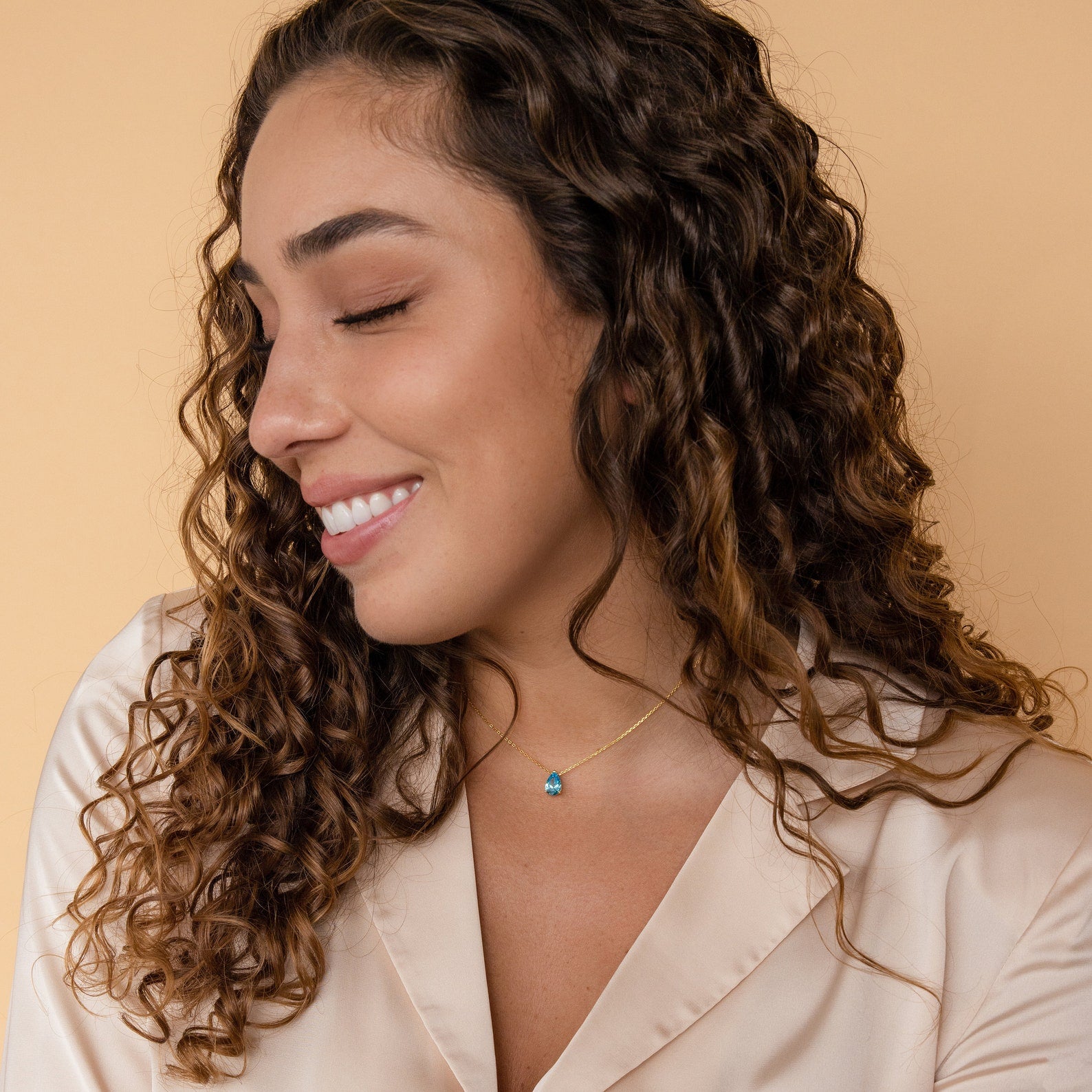 A woman with curly hair smiles, wearing a cream blouse and the Ophelia Birthstone Necklace—a delicate, minimalist piece featuring a blue pendant.