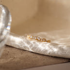 The Verity Diamond Ring with a gold cluster band and sparkling white gemstones rests on a textured, sunlit marble surface, casting delicate patterns of reflected light.