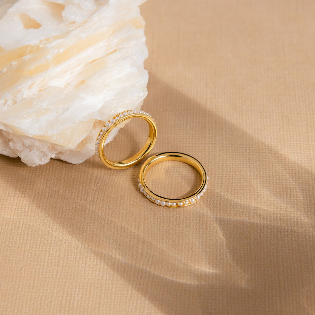 The Alicia Pearl Inlay Band, a gold stackable ring with delicate pearls, rests on a beige surface beside a large white crystal rock.