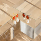 Carnelian Pearl Earrings are showcased on clear glass stands, featuring vibrant orange carnelian stones and pearl dangles for an elegant, earthy touch.