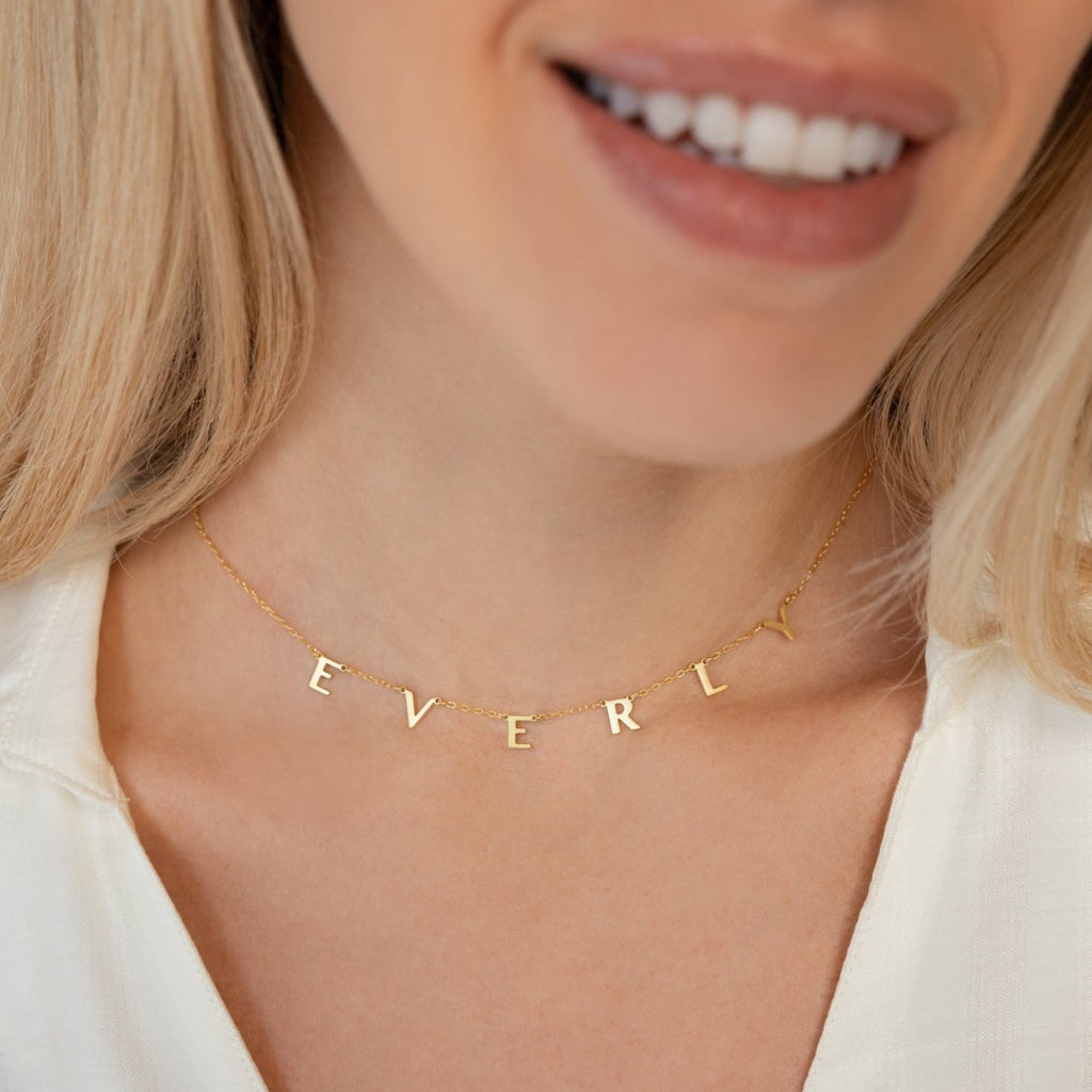 A woman in a white top smiles gently while wearing the Apollo Letter Name Necklace, a minimalist gold custom initial necklace with letter charms spelling EVERLY.