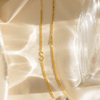 Large Letter Bracelets with S and A rest on silky cream fabric beside a glass item, ideal for pairing with your favorite everyday jewelry like a classic curb chain bracelet.
