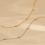 Bar Station Necklace with dainty charms and delicate chains, featuring small cylindrical beads, displayed on a beige background.