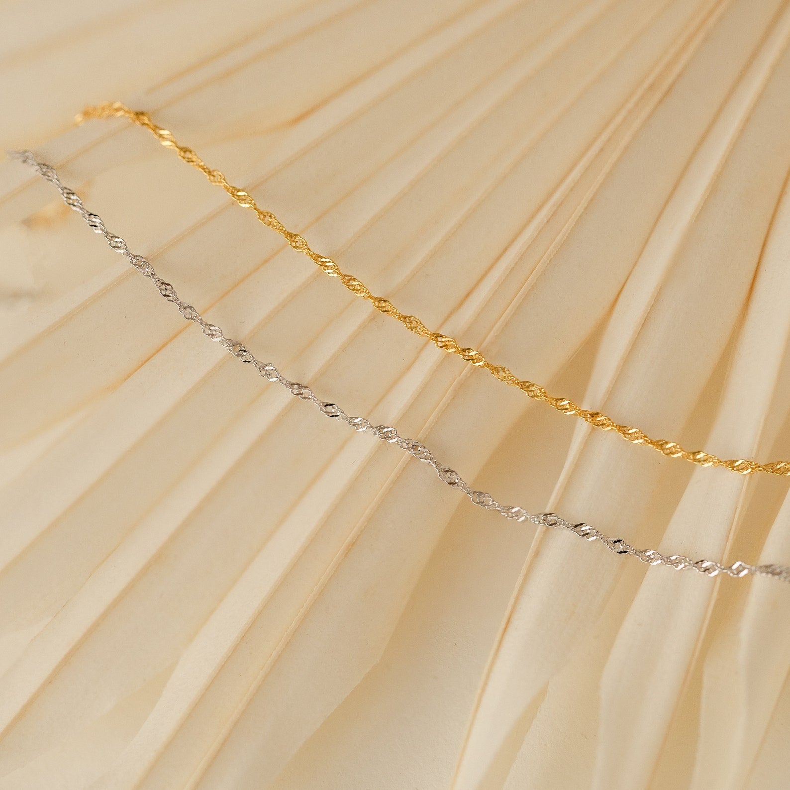 The Singapore Twist Bracelet, featuring a gold and silver twisted rope design, is displayed on a cream-colored pleated fabric background.