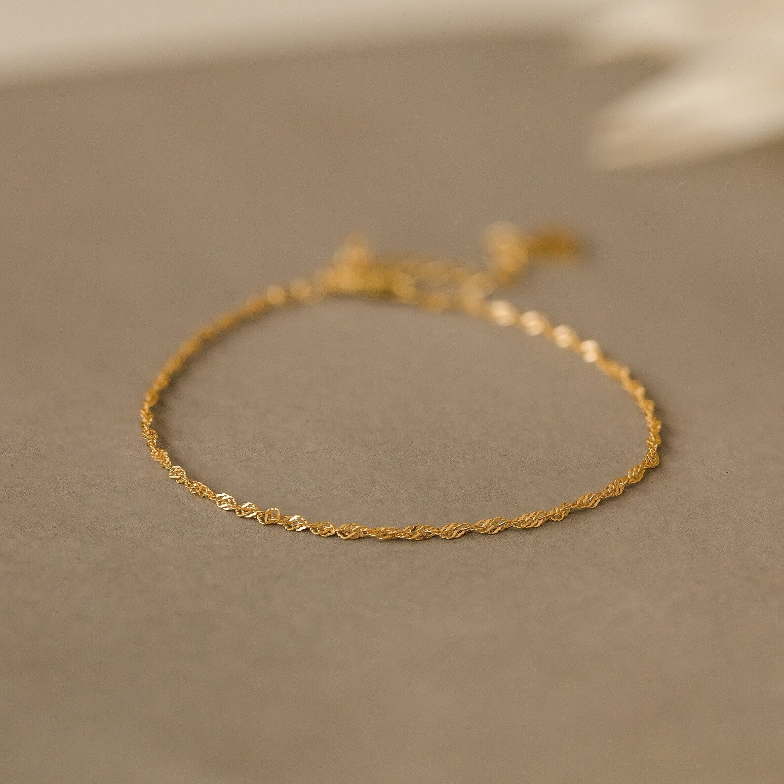 The Singapore Twist Bracelet shines on a neutral beige surface, ideal for those who appreciate dainty bracelets and minimalist elegance.