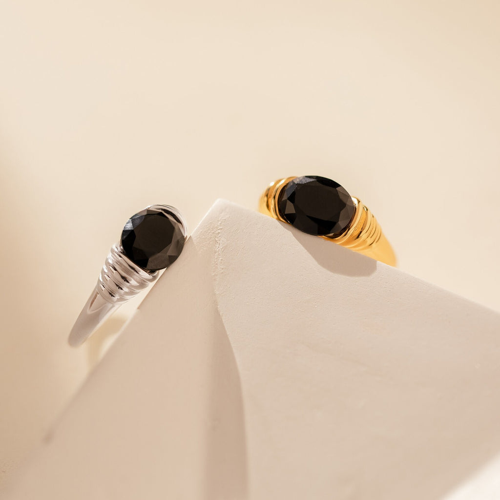 The Dusk Signet Ring, available in silver or gold with a black onyx stone, rests on a white geometric surface—ideal as statement jewelry or for adding vintage-inspired signet ring style.