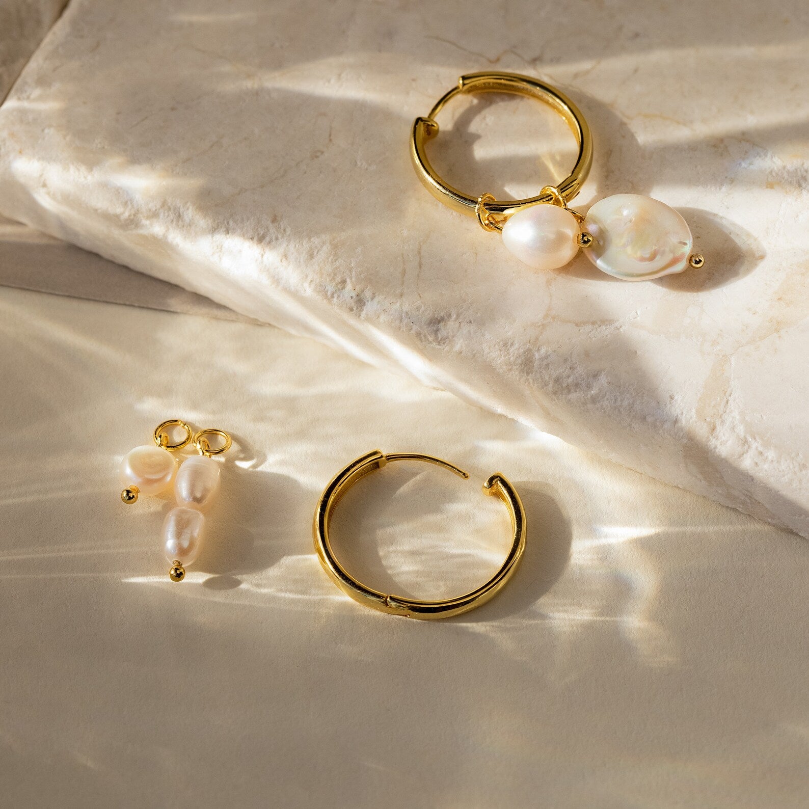 The Pearl Drop Earrings Set features gold hoops with dangling pearls, ideal for bridal wear, shown on a light stone surface under natural sunlight.
