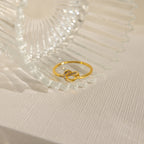 A Pave Heart Knot Ring rests against clear, textured glass on a light fabric surface—an elegant choice for a romantic gift or promise ring.