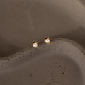 The Raindrop Pearl Onyx Studs, featuring delicate pearls and gold accents, rest on a textured brown surface, capturing timeless elegance.