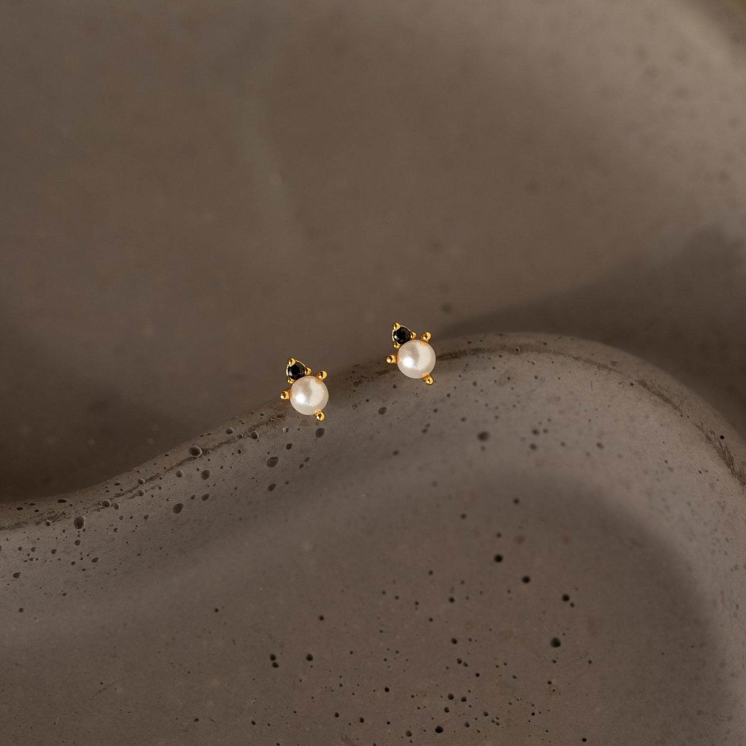 The Raindrop Pearl Onyx Studs, featuring delicate pearls and gold accents, rest on a textured brown surface, capturing timeless elegance.