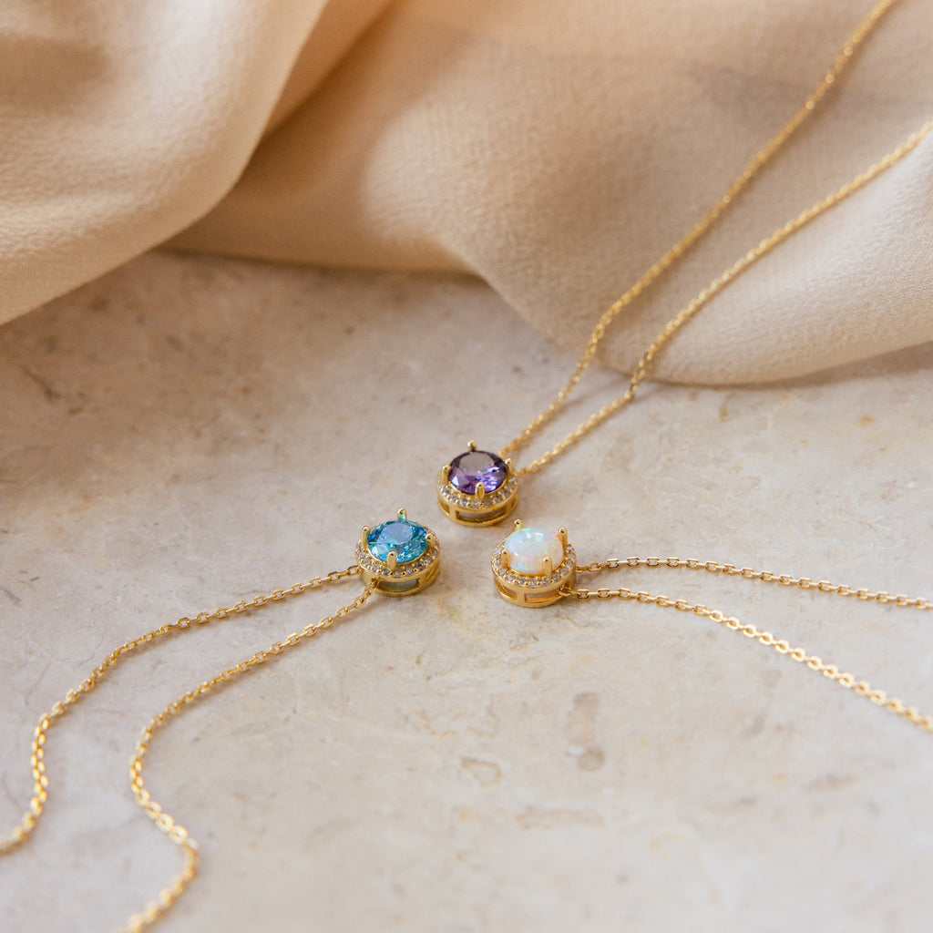Three Halo Birthstone Necklaces—featuring purple, blue, and opal gemstones—are displayed on beige fabric and stone, each providing a modern take on the classic birthstone necklace.