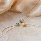 Three Halo Birthstone Necklaces—featuring purple, blue, and opal gemstones—are displayed on beige fabric and stone, each providing a modern take on the classic birthstone necklace.
