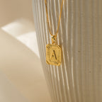 The Initial Rectangle Pendant Necklace features a gold rectangle pendant engraved with the letter A, displayed against a textured white background—an ideal personalized gift for any occasion.