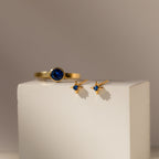Sapphire Star Studs and a gold ring with a blue stone are elegantly displayed on a white block.