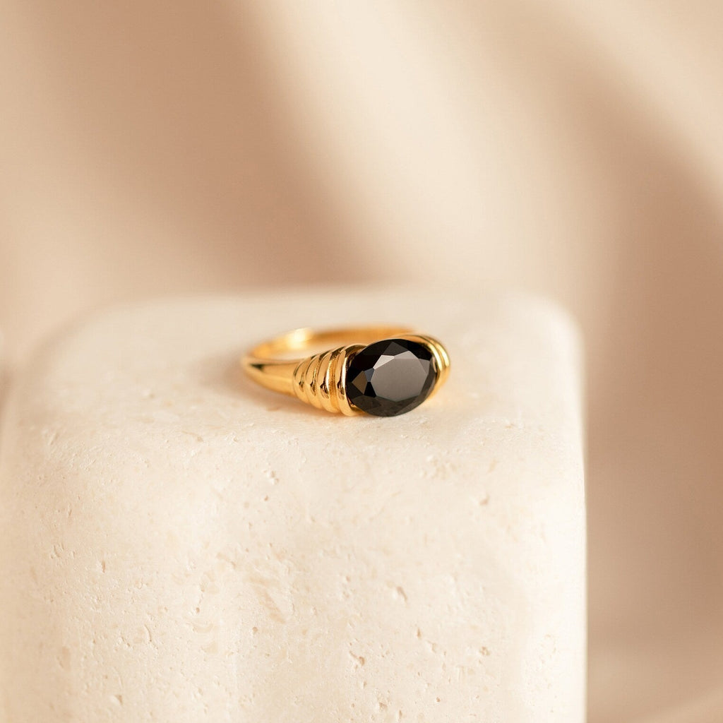 The Dusk Signet Ring features a bold black gemstone set in gold, showcased on a textured stone surface with a soft beige background.