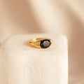 The Dusk Signet Ring features a bold black gemstone set in gold, showcased on a textured stone surface with a soft beige background.