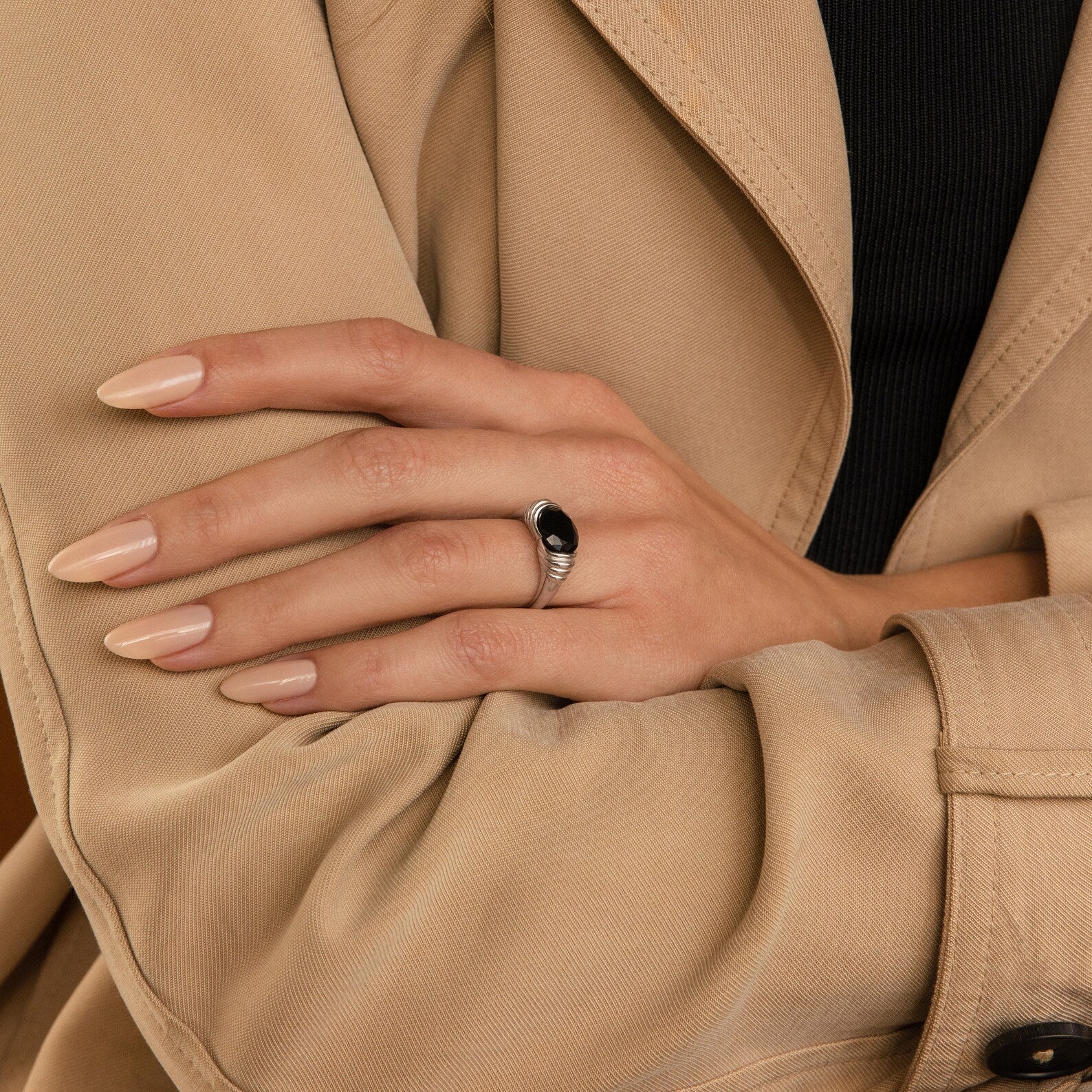 A hand with nude nails wears the Dusk Signet Ring, featuring a black stone, resting on a tan trench coat sleeve—a subtle nod to statement jewelry.