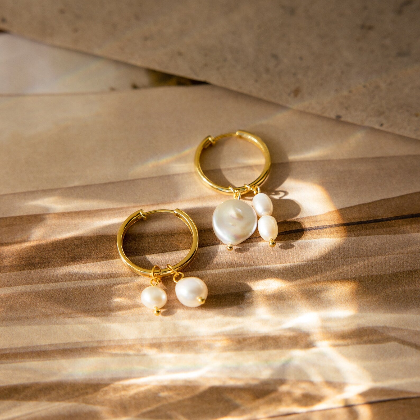 The Pearl Drop Earrings Set features gold hoops with dangling white pearls, shown shining on a sunlit, neutral-toned paper background.