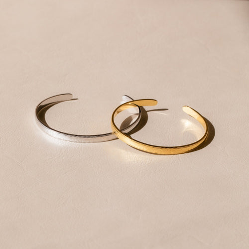 Two minimalist Alyssa Cuff Bracelets, in silver and gold, rest on a light beige textured surface—perfect to stack with your favorite pieces or wear solo for a refined look.