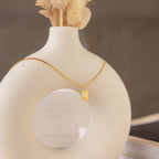 The Mini Birthstone Tag Necklace features a rectangular gold pendant with an initial, displayed on a round, cream-colored vase—a perfect personalized gift for any occasion.