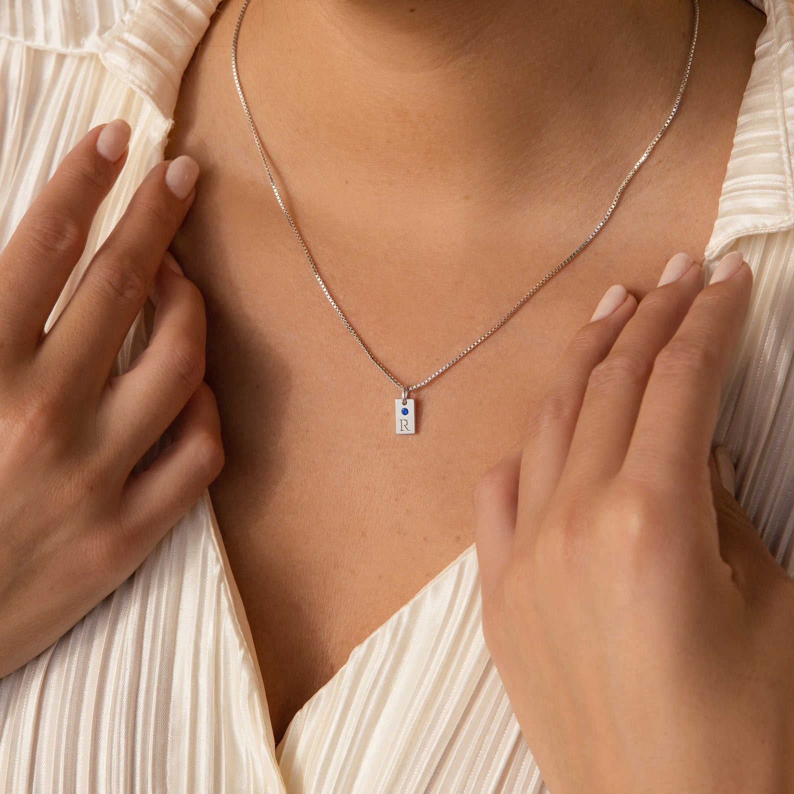 A person wearing a pleated white blouse and a Mini Birthstone Tag Necklace with a silver rectangular pendant.