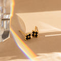 The Black Flower Studs, vintage earrings with gold and black stone accents, rest on a beige surface in sunlight, with a rainbow prism effect and a transparent crystal partially visible on the left.