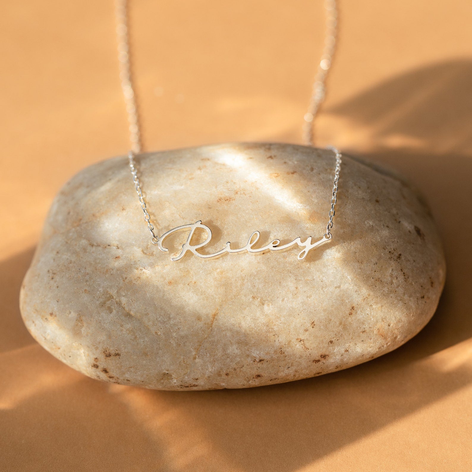 The Minimal Sideways Name Necklace features the name Riley in script font on a smooth beige stone, offering a dainty and elegant sideways design for a modern look.