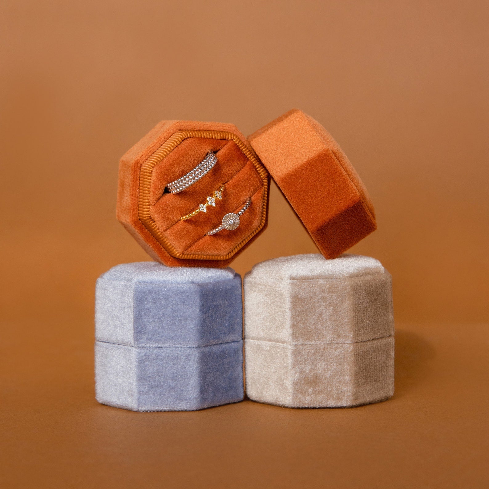 The Mystery Ring Box, a velvet jewelry case in orange, beige, and blue tones displaying rings, rests elegantly against a brown background.