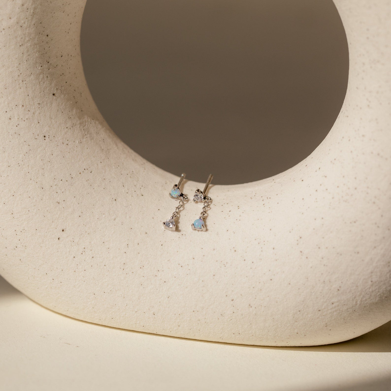 Opal Drop Studs featuring blue heart-shaped stones and delicate chain accents, showcased on a textured, cream-colored abstract sculpture.