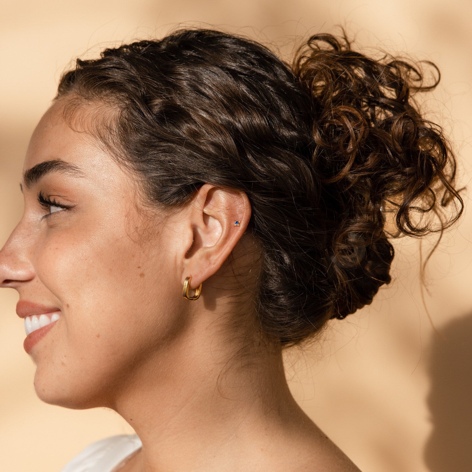 A woman with curly hair in a bun, smiling in soft natural light against a beige background, models the Sapphire Star Studs and a cartilage piercing for an effortlessly chic look.