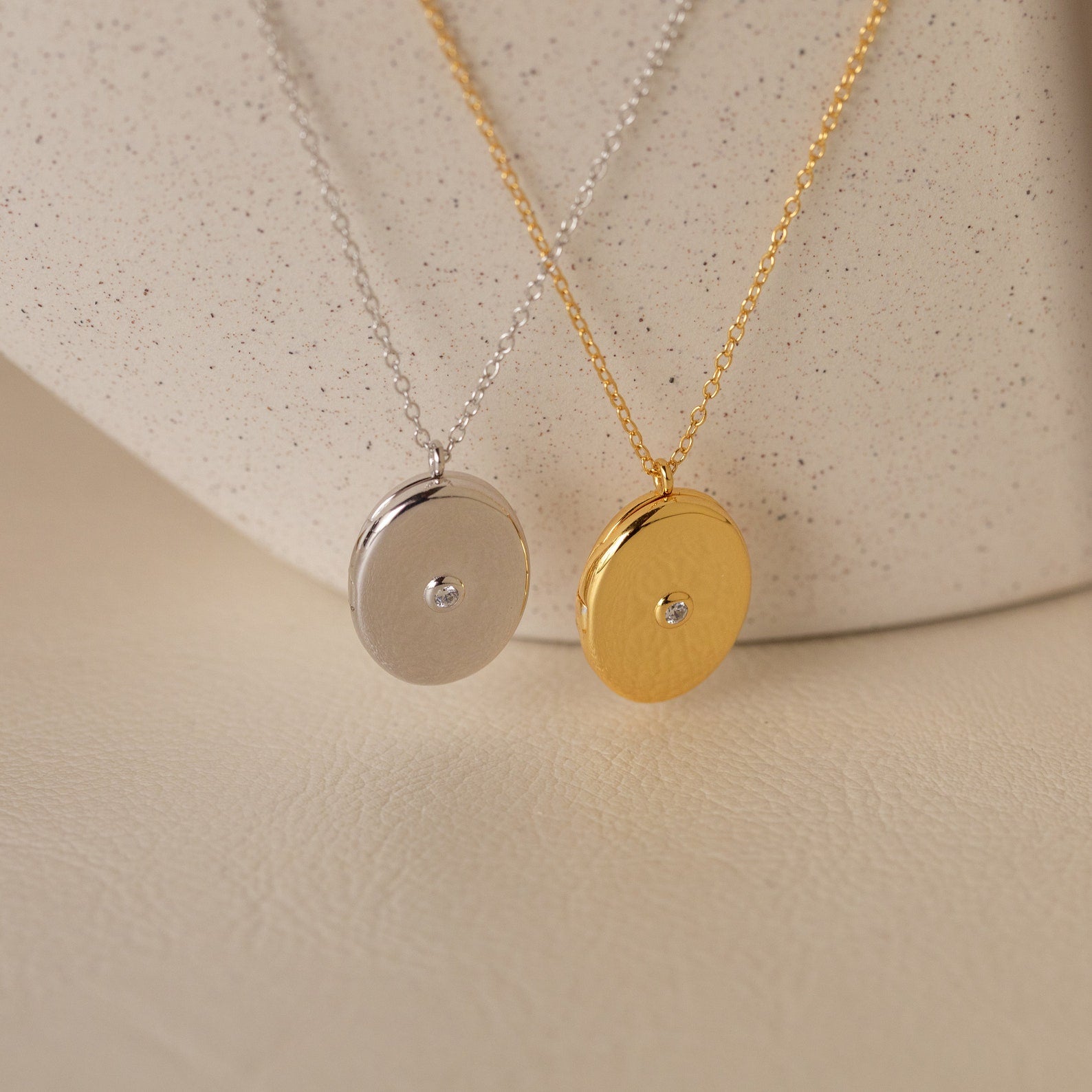 Two round pendant necklaces, one silver and one gold with a central gemstone, make a sentimental gift—perfect for pairing with the Eliza Oval Locket Necklace or a treasured photo locket. Display on a soft surface for added elegance.