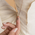 A hand displays the Mini Fingerprint Tag Necklace with a gold rectangular pendant against cream textured fabric—an elegant, personalized choice for Mother’s Day.