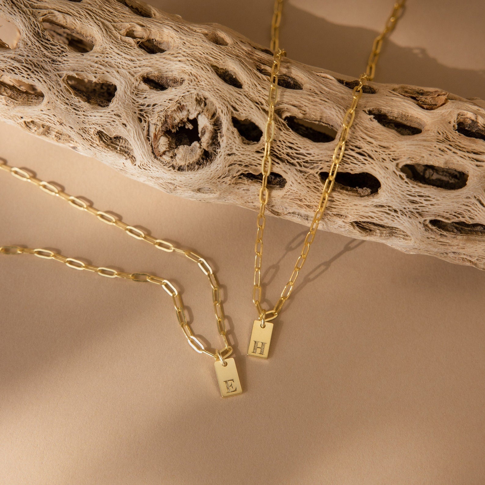 Two gold Mini Initial Paperclip Necklaces with rectangular pendants engraved with E and H rest on a textured branch—a delicate, personalized touch from the Mini Initial Paperclip Necklace collection.