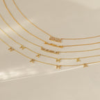 Collection of five gold “mama” necklaces displayed in a cascading layout on a smooth cream background. Designs include pave block letters, cursive script, and individual letter charms with subtle pave accents.