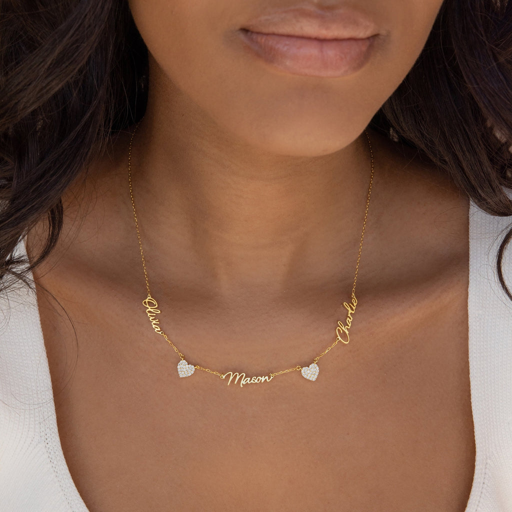 A woman wears the Grace Name Heart Necklace, featuring gold heart charms personalized in cursive with the names “Olivia,” “Mason,” and “Chloe.”.